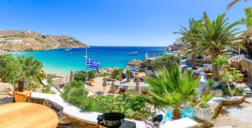 Vacanță charter Mykonos, Grecia – Plaja Paradise cu palmieri, șezlonguri și steagul Greciei.